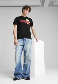 Sort t-shirt med farverigt geometrisk print, kombineret med lyseblå flagrende jeans. Modellen læner sig op ad en hvid søjle, minimalistisk baggrund.