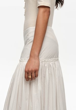 Mujer con una blusa crema de manga corta y falda blanca de talle alto con fruncidos, con un anillo dorado de varias bandas en su mano derecha.