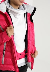 Bright pink insulated jacket with a gray hood and accents. Features a zippered pocket and a textured, puffy design. Layered with a white top.