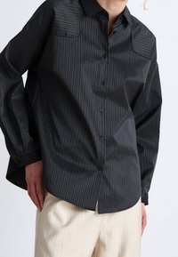 Black button-up shirt with thin vertical gold stripes, featuring a classic collar, long sleeves, and a relaxed fit with curved hem.