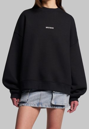 Persona che indossa una felpa nera oversize con logo bianco "WRSTBHVR" al centro, abbinata a una minigonna di jeans azzurri con dettagli a tasca.