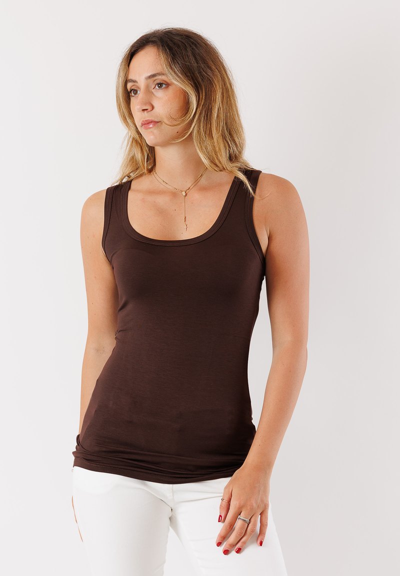 Top senza maniche marrone in morbido tessuto elasticizzato con scollatura a girocollo. Indossato con pantaloni bianchi, evidenziando una silhoutte aderente.