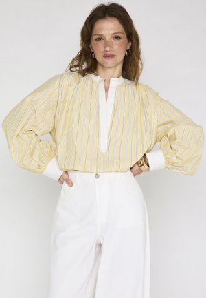 Femme portant une blouse à rayures verticales jaunes et blanches avec des manches bouffantes et un pantalon blanc taille haute, posant les mains sur les hanches.