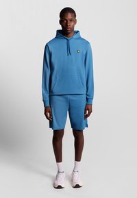 Felpa blu chiaro con tasca a marsupio e logo, abbinata a pantaloni corti coordinati. Il tessuto è liscio e ha una vestibilità comoda, adatta per un abbigliamento casual.