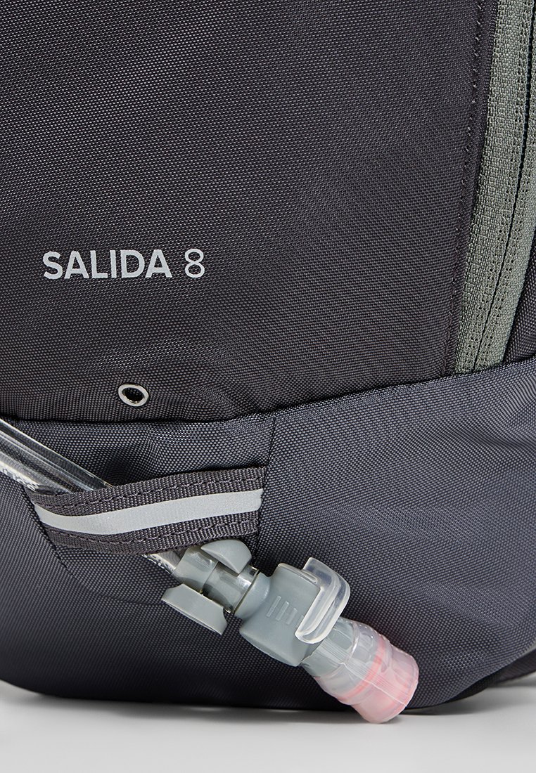 Mochila de tela negra texturizada con "SALIDA 8" impreso en blanco, con cierre de cremallera y un accesorio para tubo de hidratación en el lateral.