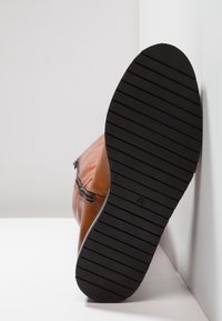 Bottine en cuir brun avec une semelle en caoutchouc rayée noire. Détail de fermeture éclair sur le côté. Le talon est arrondi et la pointe est légèrement effilée.