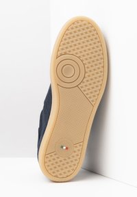Suela de zapato en goma de color marrón claro con un agarre estampado y detalle circular, unida a una parte superior de ante azul marino en vista angular sobre un fondo blanco.