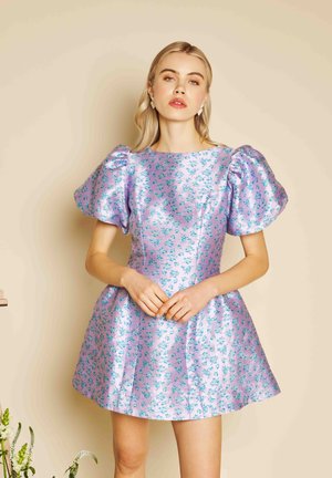 Naine seisab käed koos, seljas läikiv lavendlivärvi kleit põlvpikkuste lühikeste puffvarrukatega ja siniste lillemustritega beežil taustal.