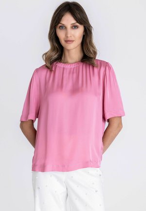 Vrouw met schouderlang bruin haar, gekleed in een losse roze blouse en witte broek met kleine versieringen, staand tegen een effen achtergrond.