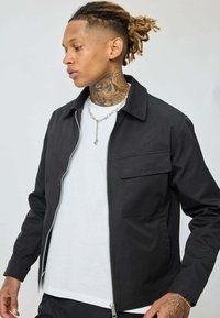 Une personne avec des dreadlocks et des tatouages sur le cou porte une veste zippée noire sur une chemise blanche, regardant sur le côté avec un collier en chaîne argentée.