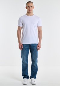 Camiseta blanca de manga corta combinada con jeans azules. El modelo está de pie, llevando zapatillas blancas. Diseño sencillo con escote redondeado.