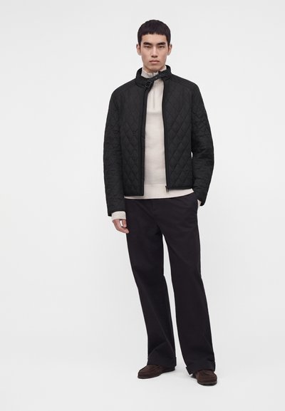 Jeune homme portant une veste matelassée noire, un pull beige clair zippé, un pantalon noir et des chaussures marron foncé, debout devant un fond uni.
