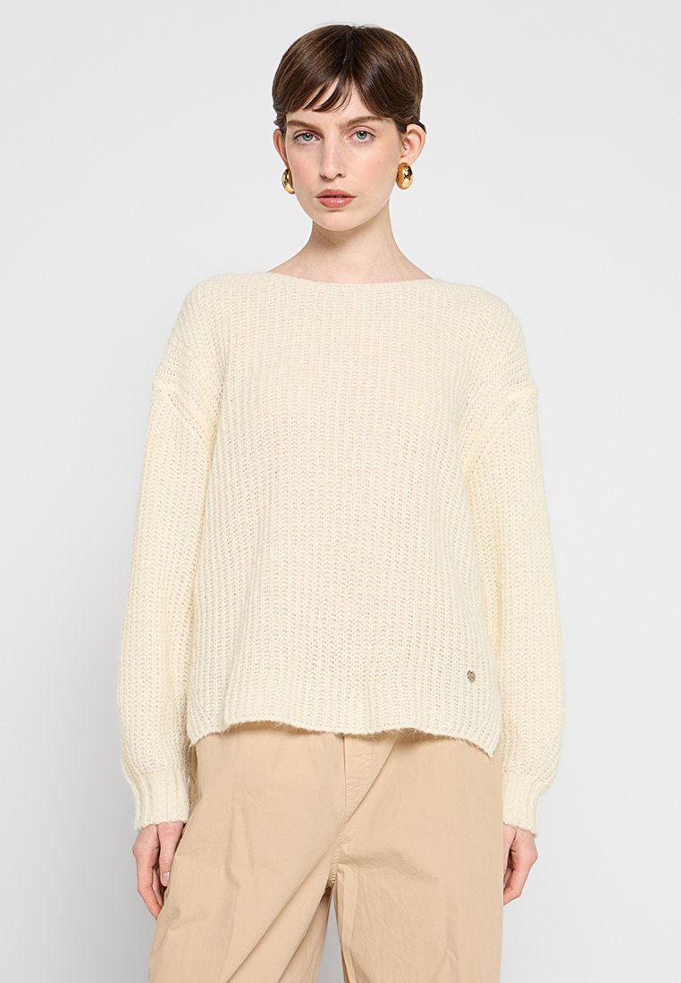 Jonge vrouw met kort bruin haar, gekleed in een crèmekleurige gebreide trui, gouden oorbellen met een ring en beige broek tegen een effen achtergrond.