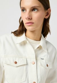 Vit denimjacka med krage, dubbla bröstfickor, kopparknappar och en liten varumärkesetikett. Har en texturerad yta.