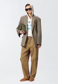 Blazer oversize marrone, felpa bianca con testo blu, pantaloni cargo beige con accenti marroni, borsa verde e occhiali da sole scuri.