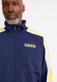 Marineblaue Adidas Trainingsjacke mit hohem Kragen, Reißverschluss, weißen und gelben Streifen an den Ärmeln und gesticktem Logo auf der Brust.