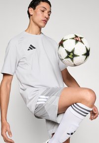 Γκρι αθλητική μπλούζα με μαύρο λογότυπο, συνδυασμένη με γκρι σορτς με ρίγες. Μία λευκή μπάλα ποδοσφαίρου με σχέδια αστεριών αιωρείται σε κίνηση.