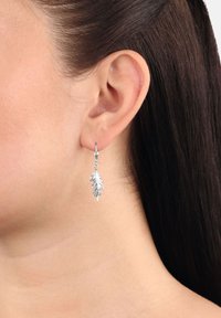 La oreja izquierda de una mujer con el cabello oscuro y liso, mostrando un arete colgante en forma de hoja plateada sobre un fondo liso.