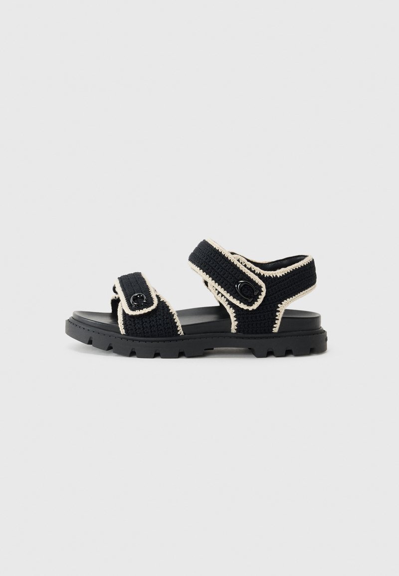 Black woven sandals with cream accents, featuring adjustable straps, a textured upper, and a rugged black rubber sole.