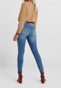 Jeans de denim azul de tiro alto con forma ajustada. Elaborados en suave algodón con un ligero desgastado y dos bolsillos traseros. Llevados con una blusa color marrón.