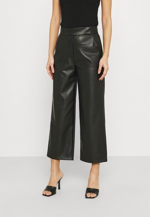 Kobieta ubrana w czarny top bez rękawów, szerokie czarne spodnie z eko skóry oraz czarne sandały na obcasie z odkrytymi palcami, stojąca na białym tle.