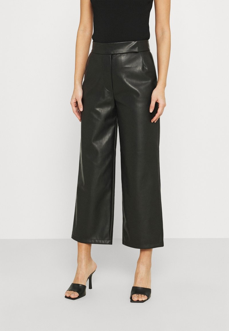 Femme portant un haut noir sans manches, un pantalon large en simili cuir noir, et des sandales à talons noires ouvertes, debout devant un fond blanc.