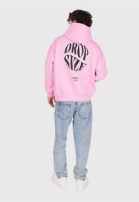 Person in pinkem Hoodie mit dem Text "DROP SIZE" auf dem Rücken, hellblauen Jeans und weißen Sneakern, steht mit dem Rücken zu einer schlichten Hintergrund.