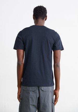 Granatowy bawełniany t-shirt z krótkimi rękawami, okrągłym dekoltem i gładką fakturą, noszony z szarymi spodniami typu cargo. Widok od tyłu.