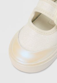 Chaussure Mary Jane blanche étincelante avec un bout rond, deux brides et une semelle extérieure lisse et brillante. Tige texturée avec des accents pailletés.