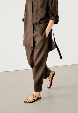 Persoon die loopt in losse bruine broek en bijpassend shirt met lange mouwen met opgerolde manchetten, zwarte sandalen met bandjes en platte zolen op een grijze vloer.