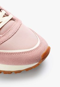 Pantofii sport roz au un superior din suede și nylon, cu o formă rotunjită, accente albe și o talpă exterioară din cauciuc texturat. Șireturile sunt de culoare crem.