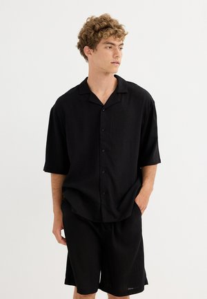 Joven con cabello rizado, vestido con una camisa negra de manga corta abotonada y pantalones cortos negros, mirando hacia su derecha con una mano en el bolsillo.