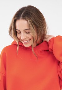 Heldere oranje hoodie met een ontspannen pasvorm, voorzien van een grote capuchon en subtiele branding bij de hals. Glad textuur en verlengde mouwen.