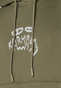 Hoodie vert olive avec un cordon de serrage et un graphique blanc représentant des mains accompagnées du mot "KARMA". Tissu texturé avec des poignets côtelés.