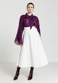Blouse en satin violet avec un détail nœud, associée à une jupe blanche évasée. Le modèle porte des bottines bordeaux brillantes. Textures lisses.
