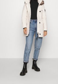 White puffer jacket with a fur-lined hood, cinched waist, and black buttons, paired with light blue jeans and black ankle boots.