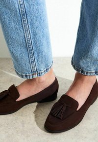 Mocasines de ante marrón con un pequeño tacón, que presentan un acento de borlas y un diseño clásico, combinados con unos vaqueros de denim ligero de color azul claro, remangados.