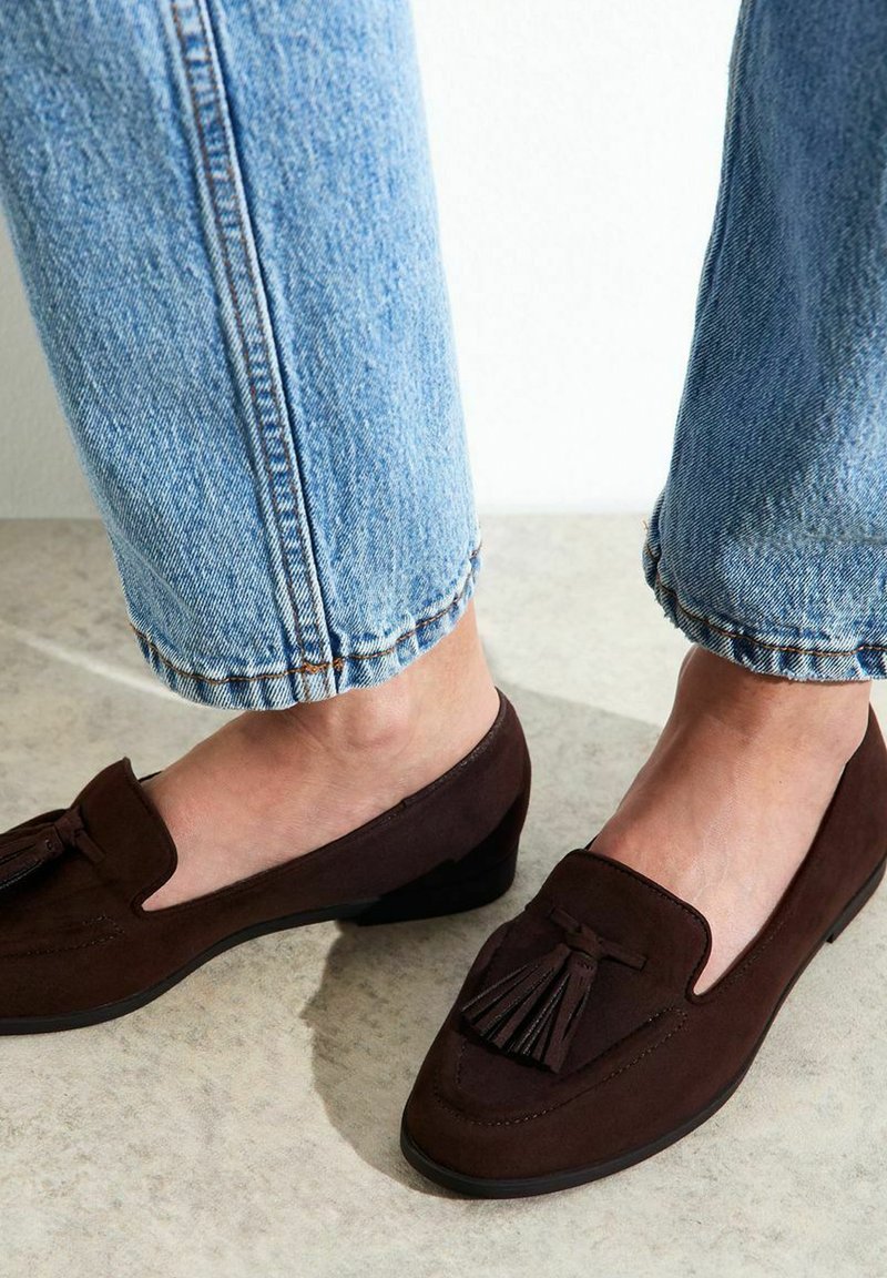 Mocasines de ante marrón con un pequeño tacón, que presentan un acento de borlas y un diseño clásico, combinados con unos vaqueros de denim ligero de color azul claro, remangados.