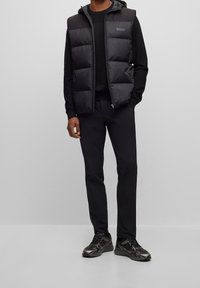 Svart puffig väst med huva, dragkedja framtill och logo-accent. Bärs över en svart tröja och kombineras med svarta byxor och mörka sneakers.