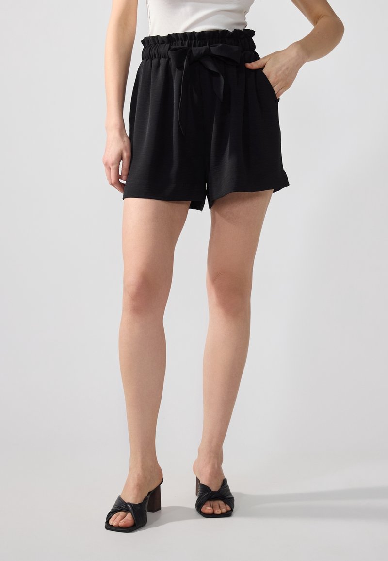 Shorts noirs avec une taille froncée, un détail de nœud noué et des poches latérales. Le tissu semble texturé et léger. Associés à des sandales à talons noires.