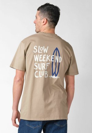 Mann trägt ein beiges T-Shirt mit weißem Aufdruck "Slow Weekend Surf Club" und blauem Surfbrett-Grafik auf der Rückseite, kombiniert mit blauen Jeans.