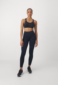 Top desportivo preto com alças ajustáveis, combinado com leggings azul-marinho de cintura alta com um padrão texturizado. Sapatos de desporto pretos.
