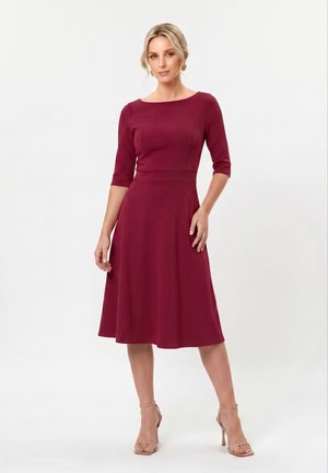 Femme aux cheveux blonds portant une robe bordeaux longueur genou avec des manches trois-quarts et des sandales à talons hauts couleur nude, debout sur un fond blanc.
