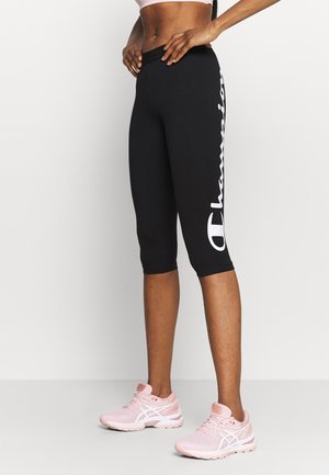 Athleta ubrany w czarne krótkie legginsy z białym napisem „Champion” oraz różowe buty do biegania, stojący na jednolitym tle.