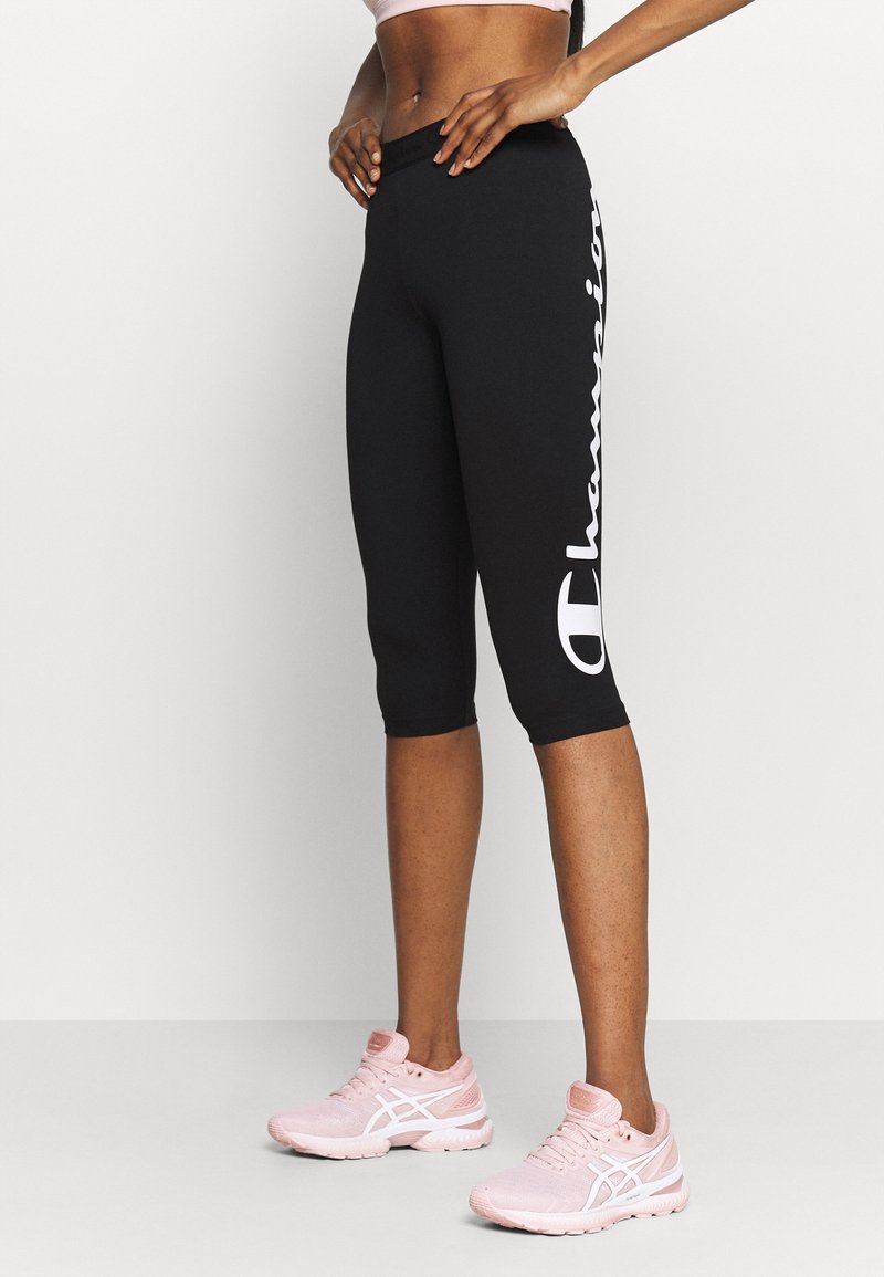 Athlète portant des leggings courts noirs avec le texte blanc « Champion » et des chaussures de course roses, debout devant un fond uni.