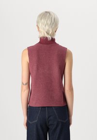 Õlavarre vabast tumepunase turtleneckiga pehme kootud top, millel on kõrge kaelus ja keha lähedane siluett, kombineeritud tumedate teksadega.