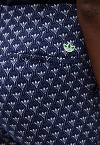 Ναυτικό μπλε ύφασμα με επαναλαμβανόμενο λευκό μοτίβο λογότυπου Adidas trefoil και ένα μικρό πράσινο και λευκό αυτοκόλλητο με το λογότυπο Adidas trefoil κοντά στην άκρη της τσέπης.