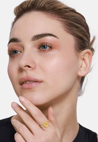 Goldring mit einem strukturierten, kreisförmigen Mittelstück, das an einer Hand getragen wird. Der Hintergrund ist schlicht, wodurch der Ring und das Gesicht des Models hervorgehoben werden.