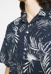 Short-sleeve navy shirt featuring a white tropical leaf pattern, button-down front, and pointed collar. Lightweight fabric with a textured look.