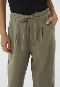 Pantalons vert olive clair avec une taille plissée et une ceinture à nouer, présentant une texture lisse et des accents en métal sur les cordons.